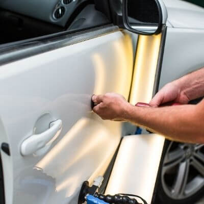 A worker using a light and a suction cup to remove a dent from a vehicle's side panel